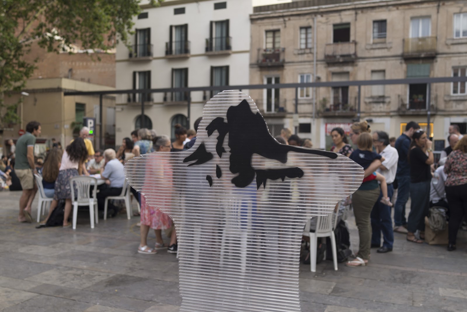 A ghost like cut out figure stuck up in the middle of a busy town square with people sat at tables and chairs in the background