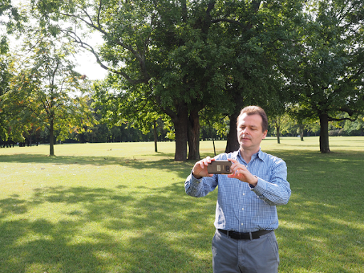 A man stands in a park with grass and trees. He is wearing a blue shirt and hold a sensor. He looks as if he is concentrating hard.