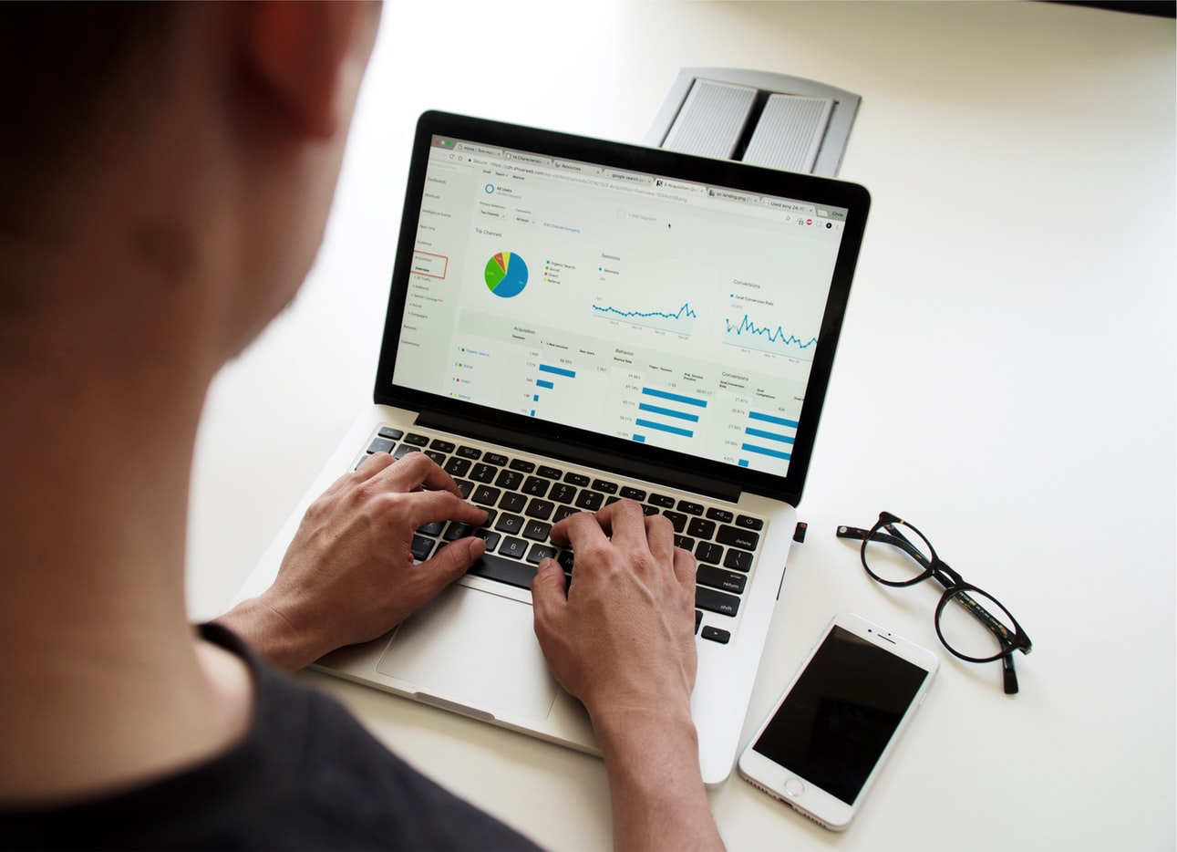 A photo taken over the shoulder of a person, probably a young white man, who is looking at a computer screen. The computer screen shows a pie chart with 4 colours and two line graphs. A pair of glasses and an iPhone are next to the keyboard, on which he is typing.