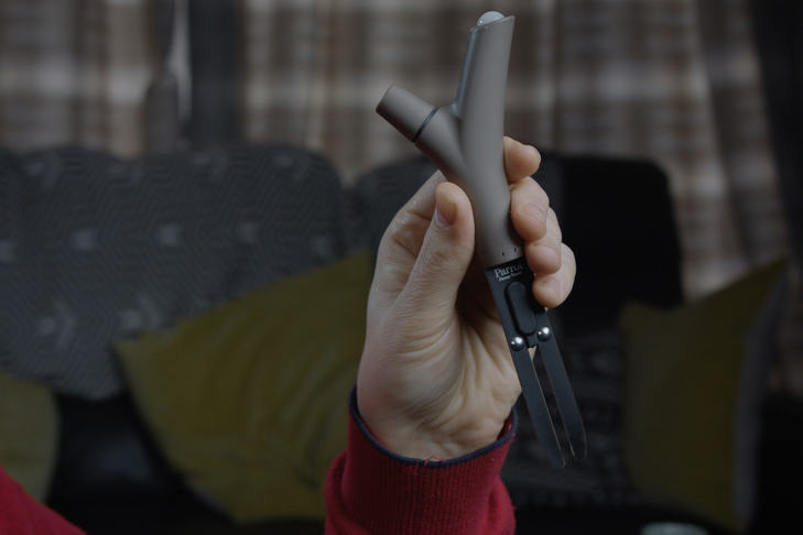 A quite dark, blurred background. In the middle is a hand (we cannot see the rest of the person, or anyone else). The hand holds a V-shaped grey plastic and black metal instrument, presumably a soil sensor.