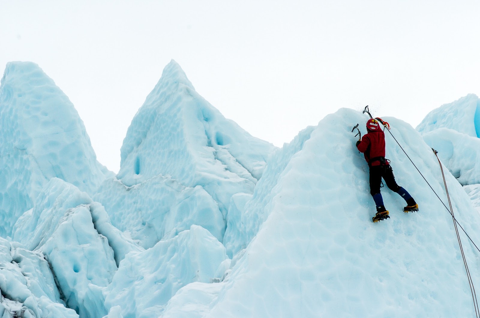 A person climbing a very steep peaked ice slope. They are dressed in burgundy gear and are very well equipped, with ropes, picks, spiked shoes, etc. Surrounding this person are several more sharp pale blue ice peaks. 