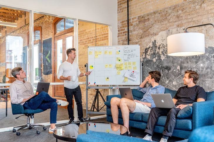 Four men in a large room with brick walls. One of them is standing by a whiteboard covered in post-it notes. He is holding a marker pen. The other men sit on comfortable furniture looking at the board. They are all in discussion.