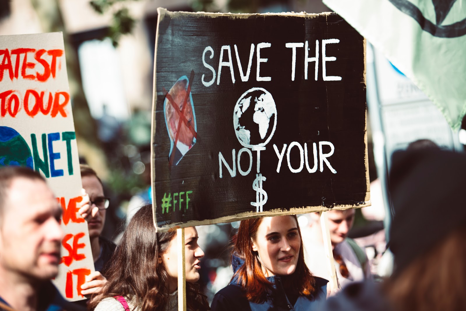 A zoomed-in photo of a protest march, focused on a black placard bearing the slogan "Save the planet not your $", with "FFF" in the corner and a picture of Bill Clinton covered by a diagonal red cross. It is held by two young white women with shoulder-length hair, one who is speaking, one who is smiling. They are surrounded by other marchers bearing similar placards.