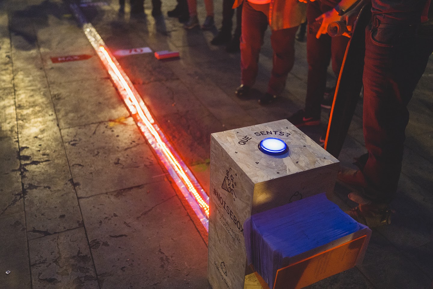 A dark street, on which many people are standing, but the image focuses on the ground so we can only see their legs and hands. In the foreground is a large box that says "Noisebox". A thin, straight, reddish line runs from the box along the street, clearly lit by a laser. On top of the box is a circular bluish light labelled "Que sents?" It seems to be a large sensor at a public event.