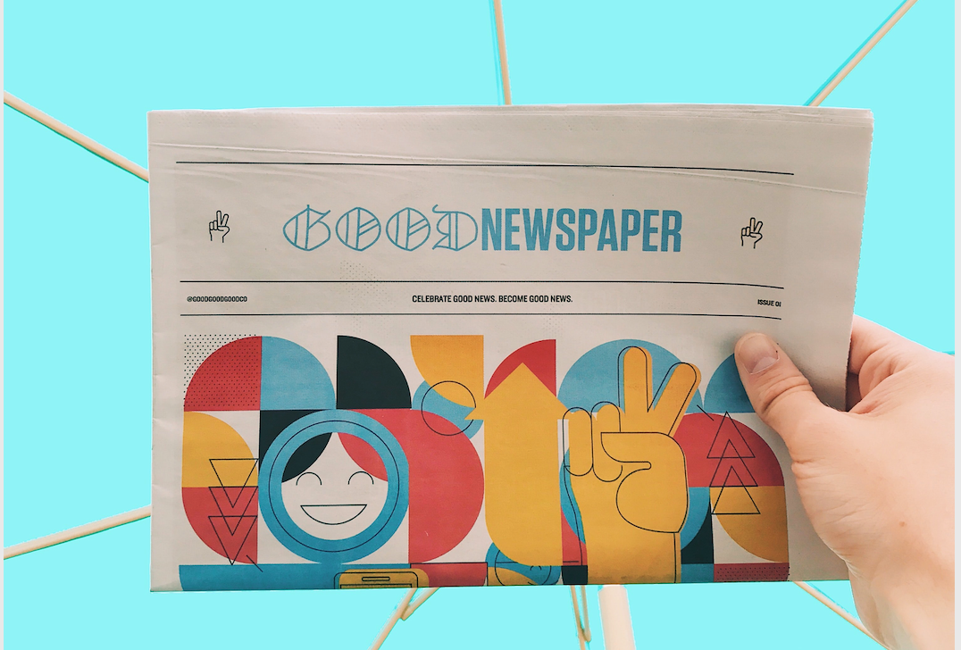 A hand of a white person holds a fiction Newspaper called Good Newspaper. Bright and cheerful colours are displayed. The background of the picture is a strong turquoise color with rays like spokes, that seem made of wood.