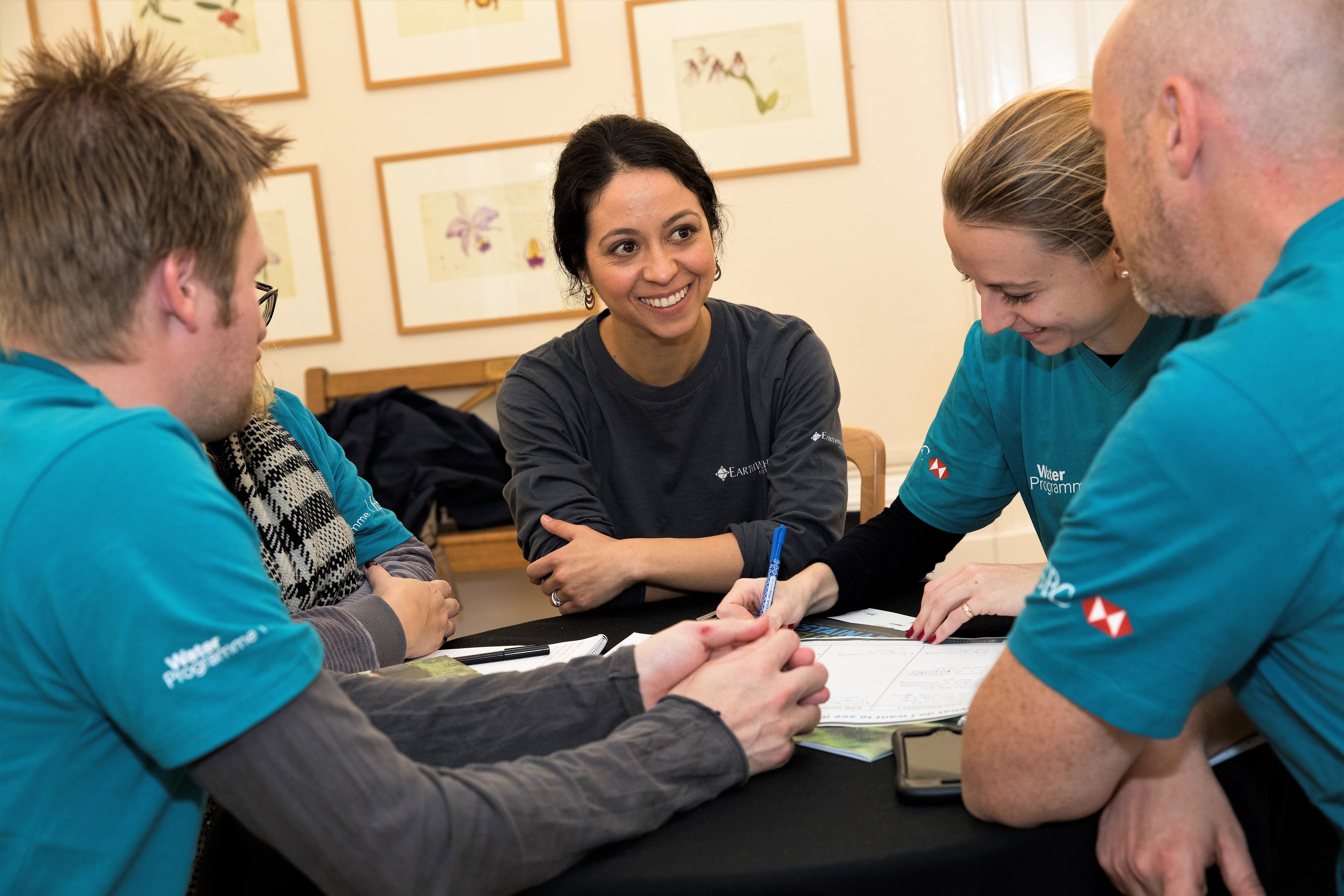 An Earthwatch employee, a smiling woman with dark hair wearing a dark blue shirt with Earthwatch's logo, is leading a training session with several people wearing blue shirts saying "Water Programme". They are all seated at one table writing on pieces of paper. 